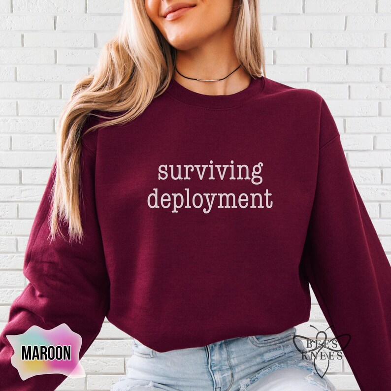 May include: Maroon sweatshirt with the text "surviving deployment" in white. The sweatshirt is a solid color and has a crew neck. The words "bees knees" are in the lower right corner and the word "maroon" is in the lower left corner.