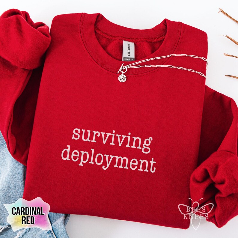 May include: A cardinal red sweatshirt with the words "surviving deployment" in white script. A silver necklace with a circular pendant rests on the sweatshirt. The words "Cardinal Red" are printed on a label.