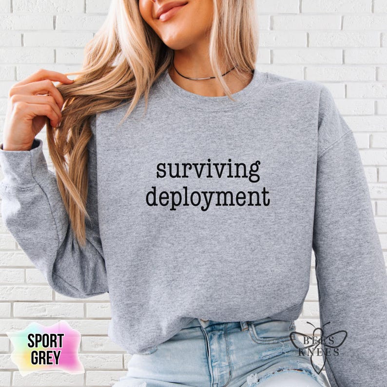 May include: Sport grey sweatshirt with the text "surviving deployment" in black. The sweatshirt is a light grey color and has long sleeves. The words "Bee's Knees" are printed on the bottom right corner.