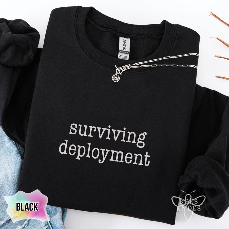 May include: Black crewneck sweatshirt with the phrase "surviving deployment" in white script. A silver necklace with a small pendant is draped across the front. The bottom right corner has the text "bees knees".