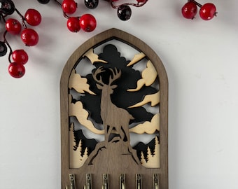 May include: A wooden key holder with a silhouette of a deer standing in a forest. The key holder has five hooks for hanging keys.