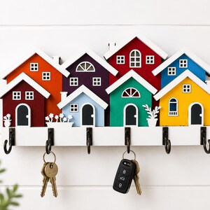 May include: A wall-mounted key holder featuring a row of seven colorful wooden houses in red, orange, purple, light blue, teal, and yellow. The houses have white roofs and black doors. Six black metal hooks are attached to the white base below.