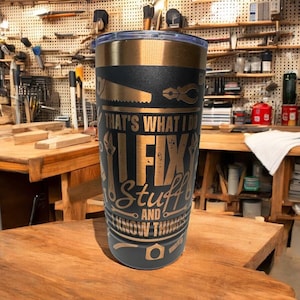 May include: A black and gold stainless steel tumbler with the text "THAT'S WHAT I DO I FIX Stuff AND KNOW THINGS." The tumbler features illustrations of tools like a saw, pliers, and wrenches. The background shows a workshop with tools.