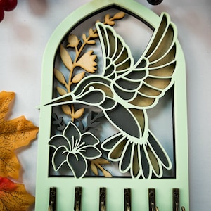 May include: A wooden key holder with a green background and a cutout design of a hummingbird and flowers. The key holder has five hooks for hanging keys.