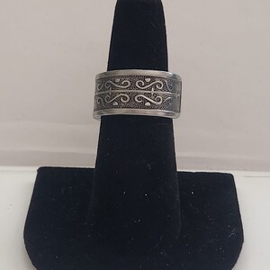 Könnte beinhalten: Ein silberner Ring mit einem dekorativen Design. Der Ring befindet sich auf einem schwarzen Ringständer.