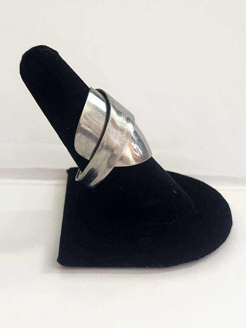 K&ouml;nnte beinhalten: Ein silberner Ring mit einem einzigartigen, gedrehten Design. Der Ring wird auf einem schwarzen Samtringhalter pr&auml;sentiert.