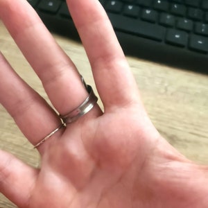K&ouml;nnte beinhalten: Ein silberner Ring mit einem breiten Band und einem strukturierten Design. Der Ring wird am Mittelfinger einer Hand getragen.