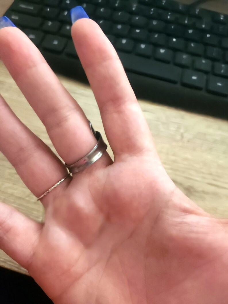 K&ouml;nnte beinhalten: Ein silberner Ring mit einem breiten Band und einem strukturierten Design. Der Ring wird am Mittelfinger einer Hand getragen.