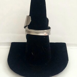 K&ouml;nnte beinhalten: Ein silberner Ring mit einem einzigartigen, offenen Design. Der Ring wird auf einem schwarzen Ringst&auml;nder pr&auml;sentiert.