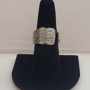 Könnte beinhalten: Ein silberner Ring mit einem geometrischen Design auf der Oberseite. Der Ring befindet sich auf einem schwarzen Ringständer.