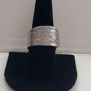 Könnte beinhalten: Ein silberner Ring mit einem floralen Design. Der Ring befindet sich auf einem schwarzen Samt-Ringständer.