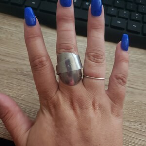 K&ouml;nnte beinhalten: Eine Hand mit blauem Nagellack tr&auml;gt einen silbernen Ring mit einem einzigartigen Design. Der Ring befindet sich am Mittelfinger und am Ringfinger befindet sich ein d&uuml;nner silberner Ring.