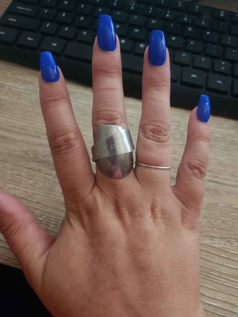 K&ouml;nnte beinhalten: Eine Hand mit blauem Nagellack tr&auml;gt einen silbernen Ring mit einem einzigartigen Design. Der Ring befindet sich am Mittelfinger und am Ringfinger befindet sich ein d&uuml;nner silberner Ring.