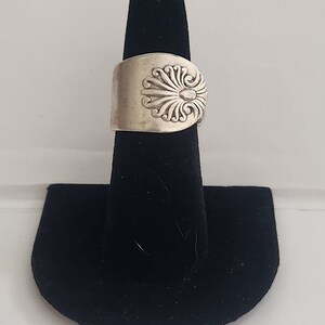 Könnte beinhalten: Ein silberner Löffelring mit einem dekorativen Design auf der Oberseite. Der Ring wird auf einem schwarzen Ringständer präsentiert.