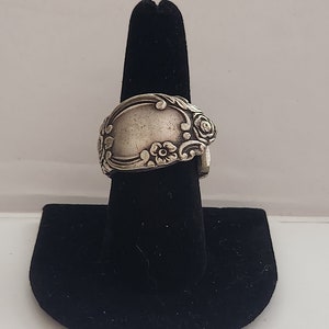 May include: A silver spoon ring with floral designs. The ring is displayed on a black ring stand.