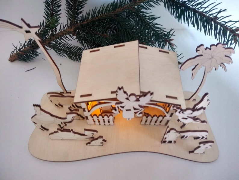 Pu&ograve; includere: Un presepe in legno con una stalla illuminata, una palma e figure di un angelo, un pastore e animali. La scena &egrave; realizzata in legno chiaro e ha un aspetto rustico e artigianale.