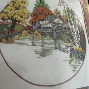 May include: An embroidery kit featuring a detailed landscape scene. The design includes a watermill, trees with autumn foliage in shades of yellow, orange, and green, and a reflective body of water. The artwork is framed by a brown border.