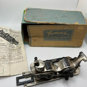 May include: A vintage "Famous Buttonhole Worker" sewing machine attachment with its original box and operating instructions. The box is tan and blue, and the machine is silver and black.