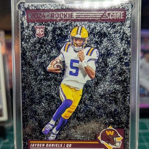 May include: A 2024 Score Rookie football card featuring Jayden Daniels in a white, yellow, and purple uniform holding a football. The card has a glittery black background, a rookie RC logo, and the text Jayden Daniels QB at the bottom.