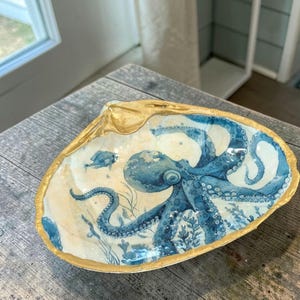May include: A decorative seashell dish with a gold rim and a blue octopus design. The octopus is surrounded by aquatic plants and a small fish. The dish is resting on a wooden surface.
