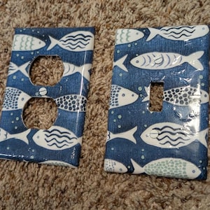 May include: Two rectangular wall plates featuring a dark blue background with a repeating pattern of white and light blue fish. One plate has two circular openings for a duplex outlet, and the other has a single rectangular opening for a light switch.