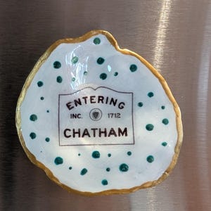 Puede incluir: Un plato de cerámica blanco, de forma irregular, con borde dorado y lunares turquesa. En el centro, el texto "ENTERING CHATHAM INC. 1712" en un marco rectangular. Probablemente un recuerdo o artículo decorativo.