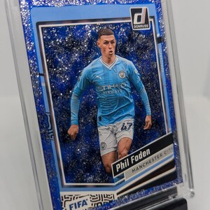May include: A Donruss trading card featuring Phil Foden of Manchester City in a light blue jersey. The card has a glittery blue border and the Donruss logo. The card is in a protective case and displayed on a black stand.