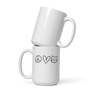 Peut inclure: Deux mugs en céramique blancs empilés. Le mug du bas présente un dessin au trait noir d'un symbole de paix, d'un cœur et d'une tête de chat. Le mug du haut est blanc uni.