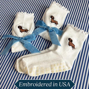 Könnte beinhalten: Weiße Socken mit Dackel-Stickerei und Rüschenkante, gebunden mit hellblauen Samtbändern. Die Socken haben rutschfeste Griffe an den Sohlen. Der Text "Embroidered in USA" befindet sich unten.