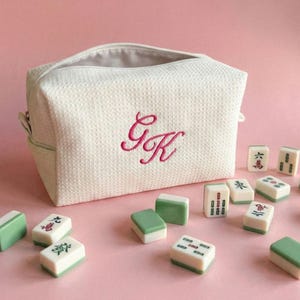 May include: A white textured cosmetic bag with a zipper closure and the embroidered letters "GK" in pink. Scattered around the bag are several green and white Mahjong tiles, creating a playful arrangement on a pink background.