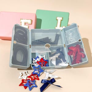Pode incluir: Uma caixa de armazenamento azul claro com vários compartimentos contendo vários acessórios de cabelo, incluindo elásticos pretos, grampos de cabelo e presilhas em forma de estrela vermelhas, brancas e azuis. Duas outras caixas são visíveis, uma rosa e outra verde, cada uma com uma letra dourada.