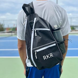 May include: A black and white tennis bag with a white zipper and a white monogram that says "RKR". The bag is worn on the back of a person wearing a white shirt and blue shorts.