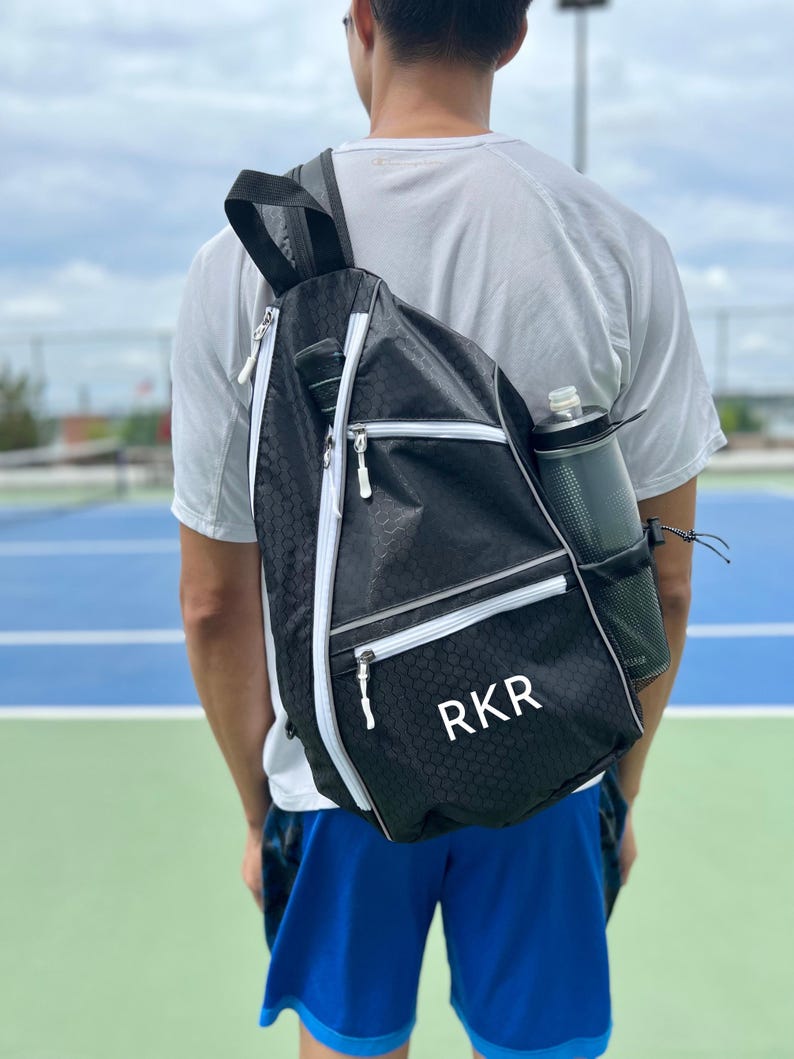 May include: A black and white tennis bag with a white zipper and a white monogram that says "RKR". The bag is worn on the back of a person wearing a white shirt and blue shorts.