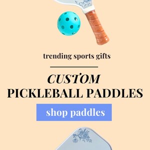 Peut inclure: Une raquette de pickleball blanche avec un motif floral bleu et le nom "Winnie" &eacute;crit dessus. La raquette est pr&eacute;sent&eacute;e dans une bo&icirc;te avec deux balles de pickleball jaunes.