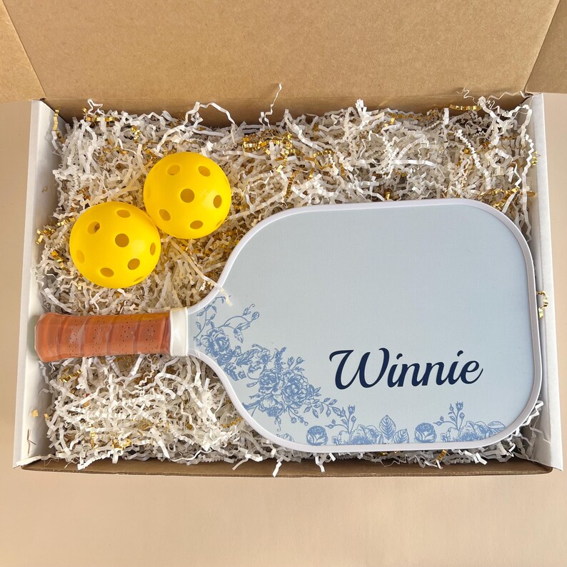 Peut inclure: Une raquette de pickleball blanche avec un motif floral bleu et le nom "Winnie" imprim&eacute; dessus. La raquette est dans une bo&icirc;te avec des balles de pickleball jaunes et du papier blanc d&eacute;chiquet&eacute;.