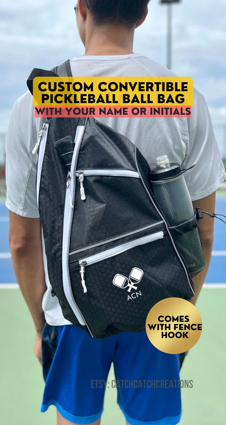 May include: A black pickleball bag with white accents and a mesh water bottle holder. The bag has a zippered compartment for paddles and a fence hook. The bag is worn by a person on a tennis court. The text on the bag reads "CUSTOM CONVERTIBLE PICKLEBALL BALL BAG WITH YOUR NAME OR INITIALS" and "ACN". The text on the gold circle reads "COMES WITH FENCE HOOK". The text at the bottom reads "ETSY: CATCHCATCHCREATIONS".