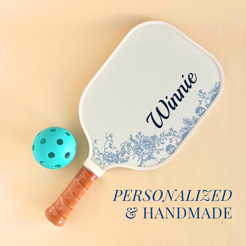 Custom pickleball paddle that has blue florals on a white background emphasizing the toile french country trend. The grip of the paddle is tan.