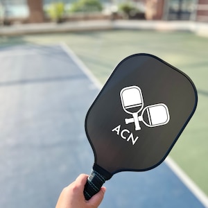 May include: A black pickleball paddle with a white logo that says "ACN" and two white pickleball paddles in the logo. The paddle is being held by a hand.
