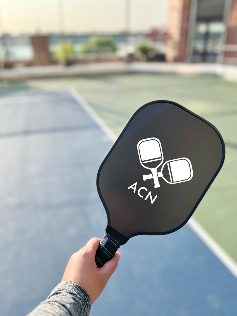 May include: A black pickleball paddle with a white logo that says "ACN" and two white pickleball paddles in the logo. The paddle is being held by a hand.