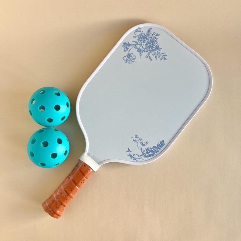 Peut inclure: Une raquette de pickleball blanche avec un motif floral bleu et une poign&eacute;e marron. Deux balles de pickleball turquoise sont au premier plan.