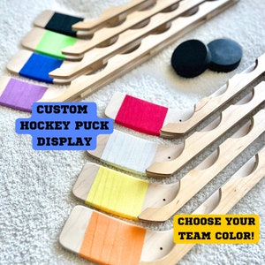 Puede incluir: Expositor de palos de hockey de madera con rayas de varios colores. El texto "CUSTOM HOCKEY PUCK DISPLAY" est&aacute; en la esquina superior izquierda. El texto "CHOOSE YOUR TEAM COLOR!" est&aacute; en la esquina inferior derecha.