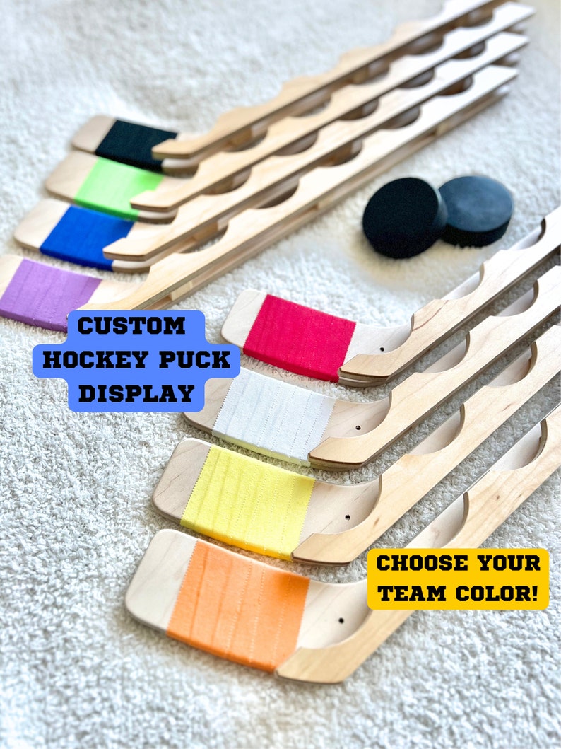 Puede incluir: Expositor de palos de hockey de madera con rayas de varios colores. El texto "CUSTOM HOCKEY PUCK DISPLAY" est&aacute; en la esquina superior izquierda. El texto "CHOOSE YOUR TEAM COLOR!" est&aacute; en la esquina inferior derecha.