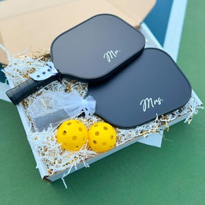 May include: A white box filled with gold and white shredded paper. Two black pickleball paddles with the words "Mr." and "Mrs." written in white are in the box. Two yellow pickleball balls are also in the box.