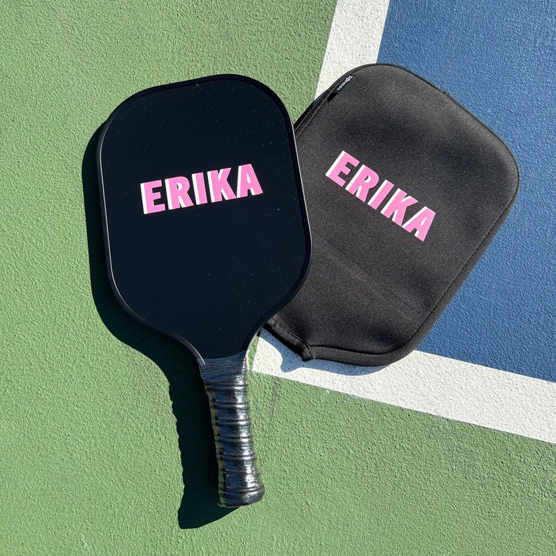 May include: A black paddleball racket with a black handle and the name "ERIKA" printed in pink letters. A black carrying case with the same name printed in pink letters is next to the racket.