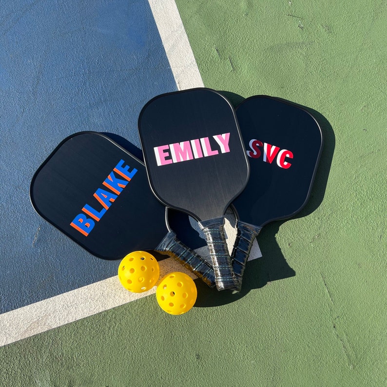 May include: Three black paddleball rackets with the names "Blake", "Emily", and "SVC" printed on them. Two yellow paddleballs are in front of the rackets.