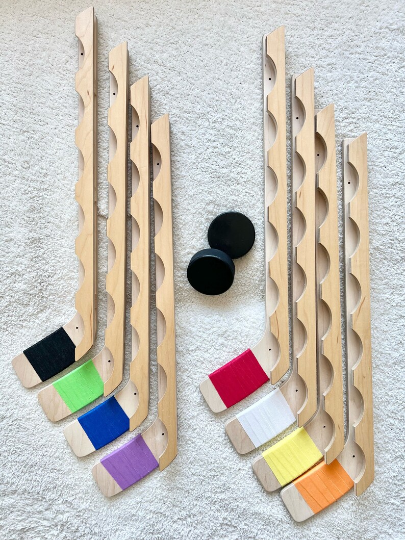 Puede incluir: Conjunto de 10 palos de hockey de madera con cinta de colores en las palas. Los palos est&aacute;n dispuestos en dos filas, la fila superior tiene cinta negra, verde, azul y morada, y la fila inferior tiene cinta roja, blanca, amarilla y naranja.