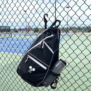 May include: A black and white sports bag with a white zipper and a white logo that says "ACN" and two white paddles. The bag is hanging on a chain link fence with a water bottle in the side pocket.