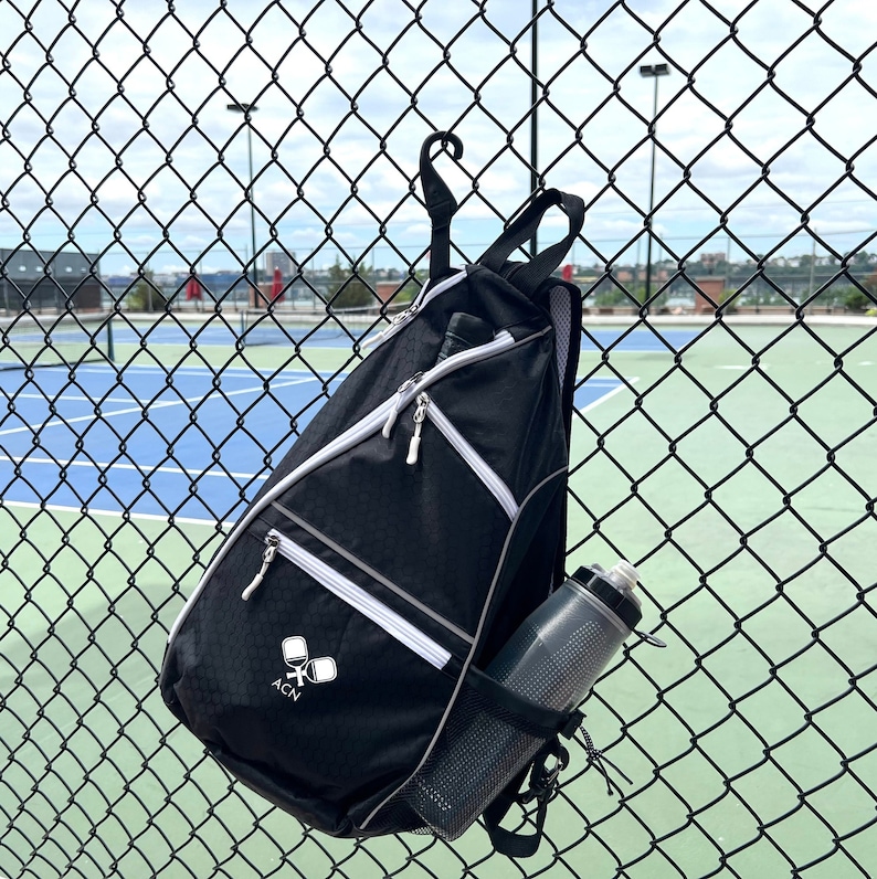 May include: A black and white sports bag with a white zipper and a white logo that says "ACN" and two white paddles. The bag is hanging on a chain link fence with a water bottle in the side pocket.