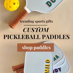Pu&ograve; includere: Quattro racchette da pickleball bianche con manici marroni. Le racchette sono personalizzate con i nomi Donna, Kody, Milo e Donna. Una palla da pickleball gialla &egrave; in primo piano. Il testo "trending sports gifts CUSTOM PICKLEBALL PADDLES shop paddles" &egrave; sull'immagine. Il testo "ETSTY: @catchcatchcreations" &egrave; sull'immagine.