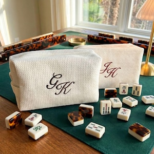 May include: Two off-white, textured cosmetic bags with embroidered initials, alongside a set of Mahjong tiles. The tiles are white with black and green markings, and some have a tortoiseshell pattern. The game is set on a green surface.